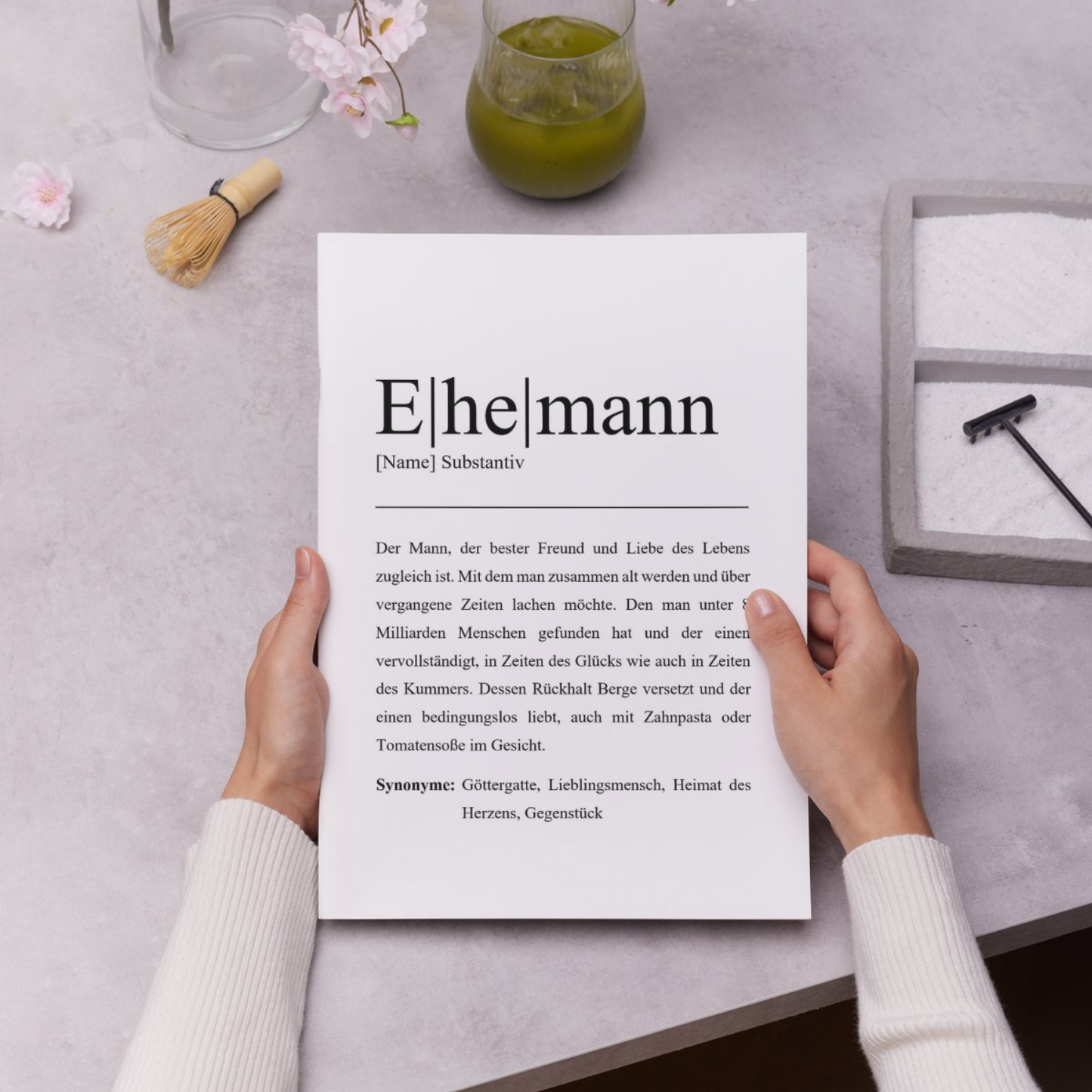 Personalisierbare Wandkunst mit Definition von Ehemann. Zeigt Hände die ein Poster halten. Ideal als Ehemann Geschenk. Dekorative Elemente wie Holzoberfläche und Matcha-Tee im Hintergrund für stilvolle Präsentation.