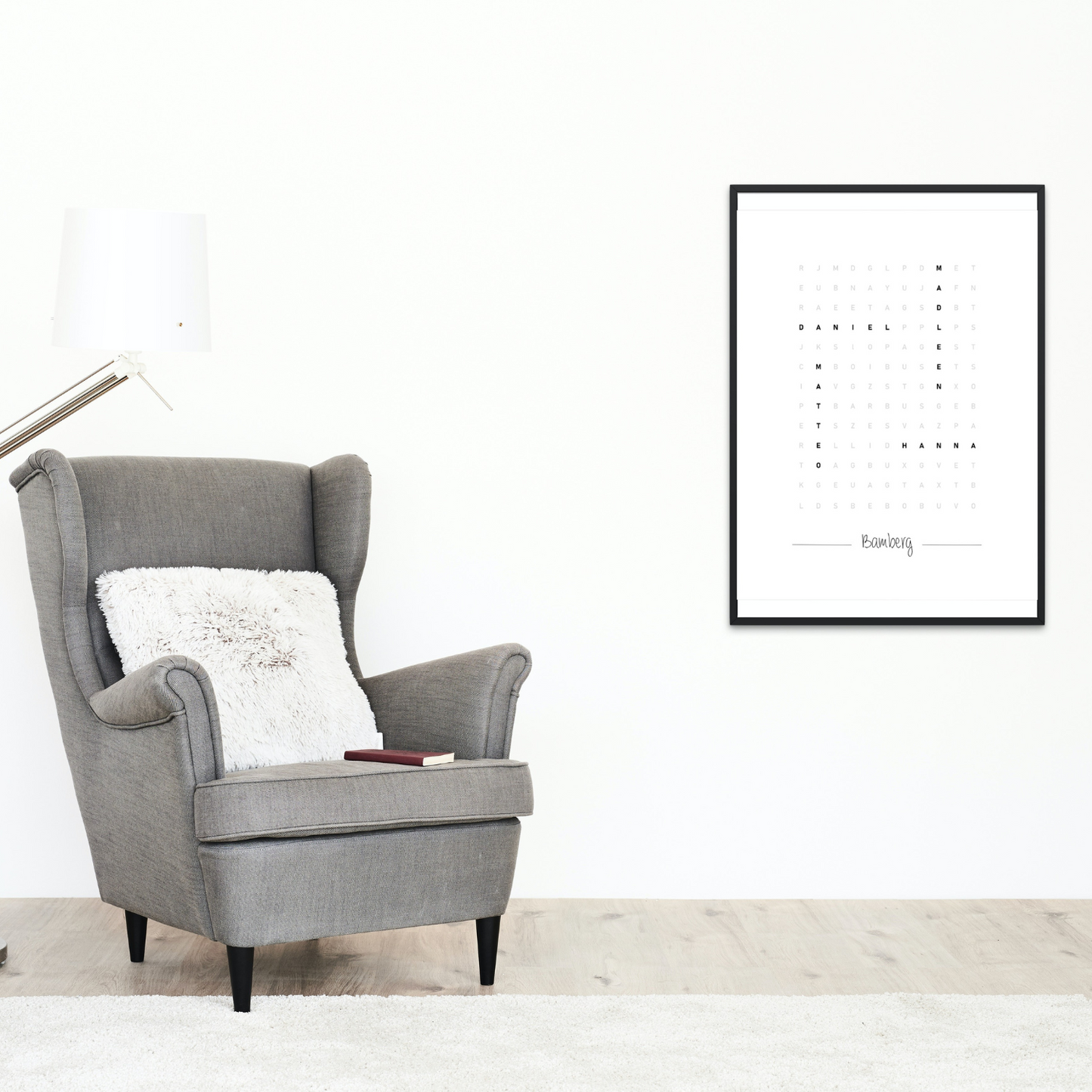 Minimalistisches, personalisiertes Wortsuche-Wandbild in schwarzem Rahmen. Perfekt für moderne Wohnzimmergestaltung. Abgebildet neben einem gemütlichen grauen Sessel mit Kissen und Buch.