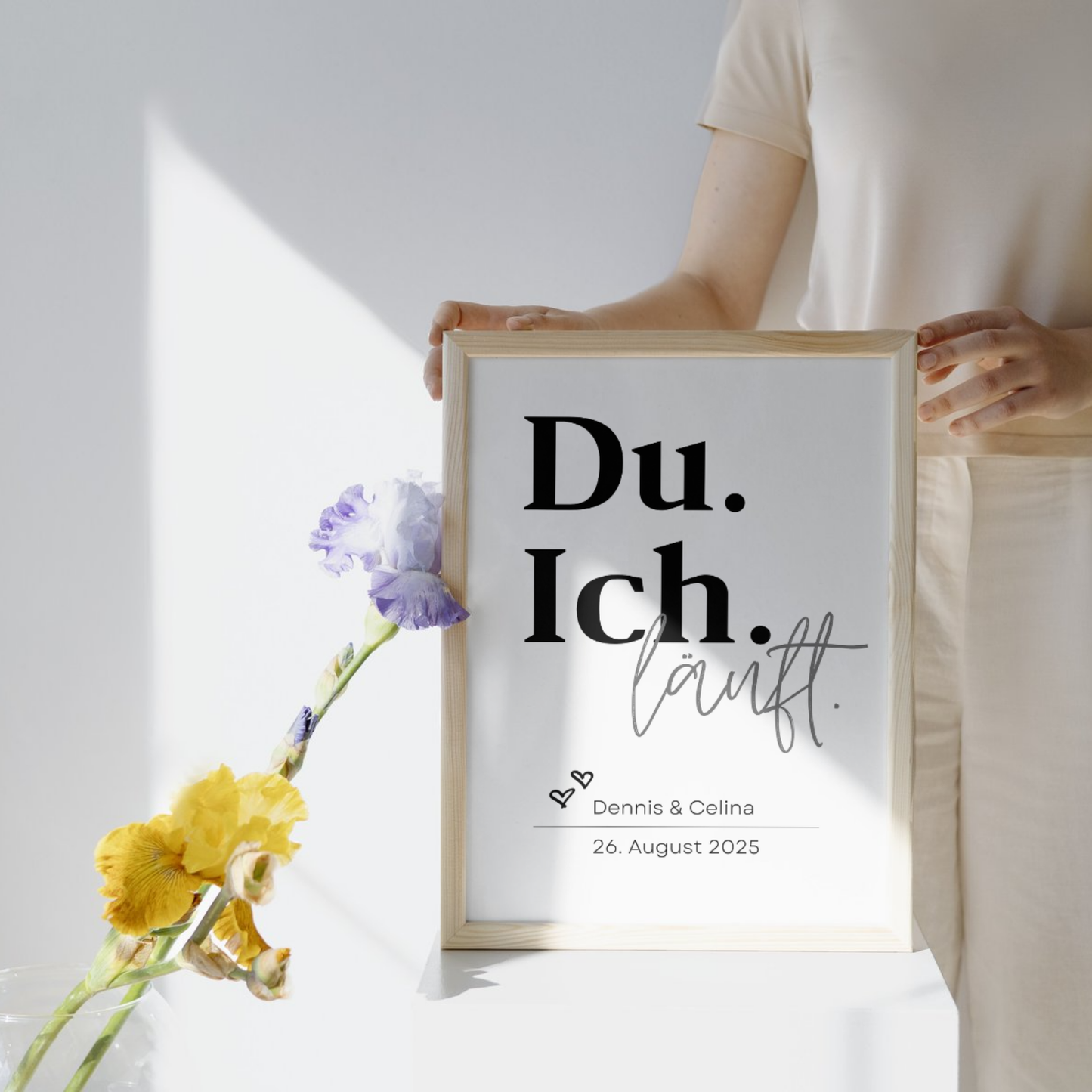 Person hält personalisiertes Poster mit dem Text Du Ich läuft in einem Holzrahmen. Entworfen für Dennis und Celina mit Datum 26 August 2025. Ideal als individuelles Geschenk für Paare. Hintergrund mit Blumen dekoriert.